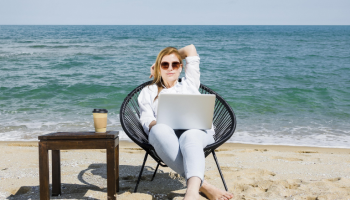 A freelancer girl working a truly remote job using her laptop sitting on a beach with sea behind her and a cup of coffee
