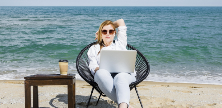 A freelancer girl working a truly remote job using her laptop sitting on a beach with sea behind her and a cup of coffee