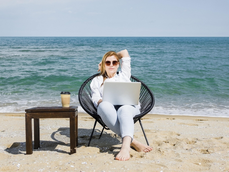 A freelancer girl working a truly remote job using her laptop sitting on a beach with sea behind her and a cup of coffee
