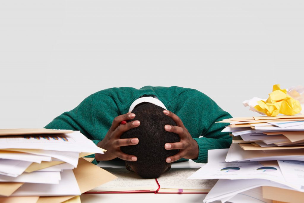 A young employee trying to streamline the work week with frustration with his head on a pile of paperwork and using Trello and Easynote as help.