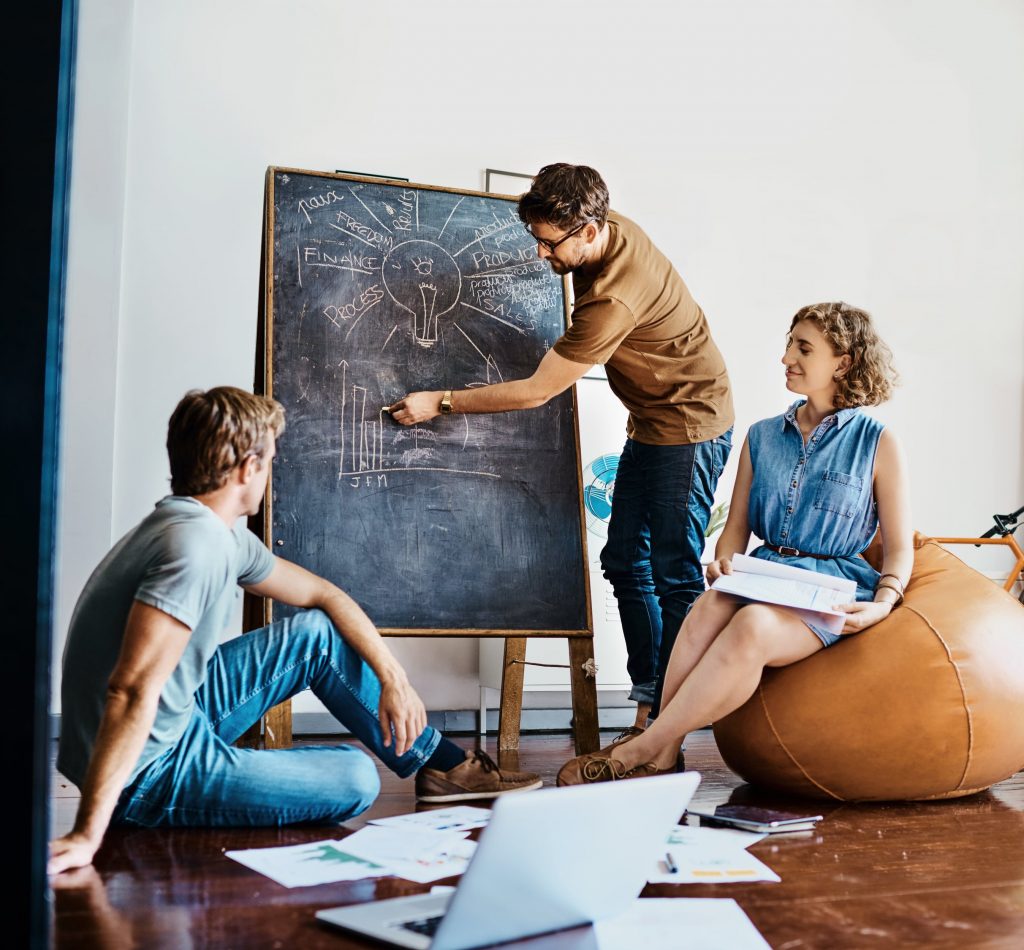 A scrum master using techniques and writing on a whiteboard to coach the team that is sitting on the floor around him in an office